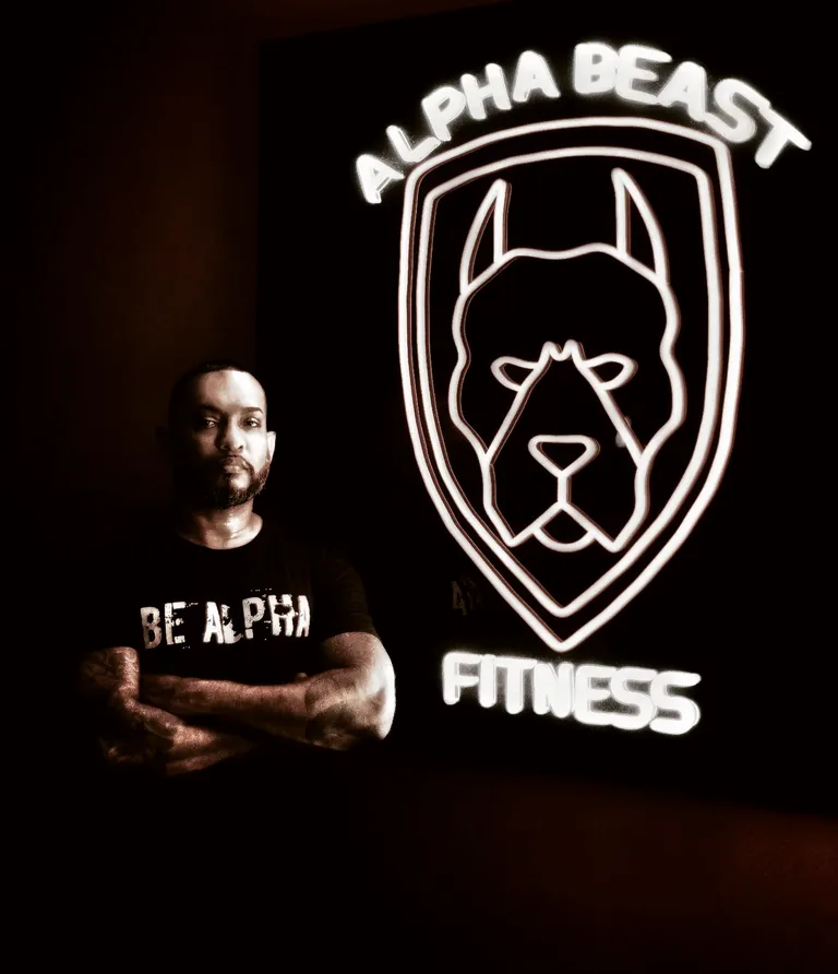 EJ Philip standing with his arms folded beside the illuminated Alpha Beast Fitness sign in the studio